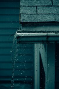 Rainwater flows from a shingled roof into clean gutters and through a downspout away from the house.